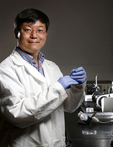 Jaeyoun (Jay) Kim holds a syringe with a micro-tentacle attached. Behind Kim is the system of nozzles, micro-manipulators and microscopic cameras developed by his research group to shape soft, rubbery PDMS material.