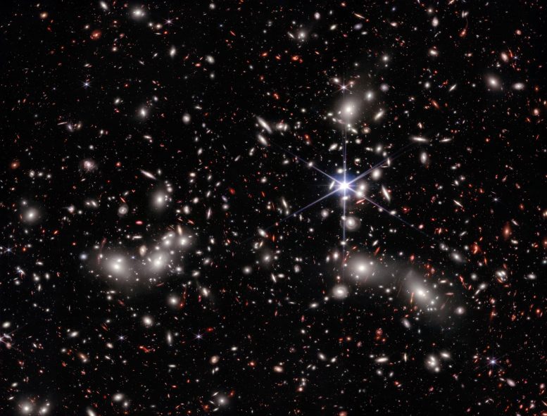 Os astrônomos estimam que aproximadamente 50.000 fontes de luz infravermelha estão representadas nesta imagem obtida pelo Telescópio Espacial James Webb, da NASA. A trajetória percorrida por essa luz abrange distâncias variadas até alcançar os detectores do telescópio, capturando a imensidão do espaço em uma única imagem. Uma estrela localizada em primeiro plano, pertencente à nossa própria galáxia e posicionada à direita do centro da imagem, exibe os distintos picos de difração do Webb. As fontes brilhantes de cor branca, envoltas por um brilho nebuloso, correspondem às galáxias presentes no Aglomerado de Pandora, um conglomerado composto por aglomerados maciços de galáxias que se encontram em processo de fusão, formando um megaaglomerado. A concentração de massa é tão intensa que o tecido do espaço-tempo é deformado pela gravidade, resultando em um efeito que desperta especial interesse para os astrônomos: uma região de lente gravitacional natural.Essas fontes, afetadas pela lente gravitacional, são visíveis na imagem na cor vermelha e frequentemente se apresentam como arcos alongados e distorcidos. Muitas dessas fontes são galáxias primitivas, cujos conteúdos são ampliados e estendidos, permitindo seu estudo detalhado pelos astrônomos. Outras fontes vermelhas presentes na imagem ainda aguardam confirmação por meio de observações complementares realizadas com o instrumento Near-Infrared Spectrograph (NIRSpec) do Webb, a fim de determinar sua verdadeira natureza. Um exemplo intrigante é uma fonte extremamente compacta, visualizada como um pequeno ponto vermelho, mesmo sob a ampliação proporcionada pela lente gravitacional. Uma possibilidade é que esse ponto represente um buraco negro supermassivo existente no início do universo. Os dados obtidos pelo NIRSpec fornecerão medições de distância e detalhes sobre a composição de fontes selecionadas, proporcionando uma riqueza de informações anteriormente inacessíveis acerca do universo e de sua evolução ao longo do tempo.